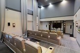This private screening room is thoughtfully designed to replicate the atmosphere of a professional movie theater. The space features tiered, stadium-style seating arranged across multiple rows, ensuring unobstructed sightlines to the large flat-screen display mounted at the front of the room. Each seat is upholstered in soft, neutral-toned leather and equipped with built-in cup holders, prioritizing comfort during extended viewing sessions.

The room's soaring ceilings add a dramatic sense of volume and grandeur, while floor-to-ceiling drapes in a light, neutral fabric line the walls, contributing to both the aesthetic and acoustic quality of the space. Subtle wall-mounted sconces provide ambient lighting that can be dimmed to enhance the cinematic experience. The walls are finished in a sophisticated two-tone gray palette, with architectural paneling details that add texture and visual interest.

The flooring features wide-plank wood-tone material that complements the overall refined design scheme. Recessed ceiling lighting offers precise illumination control, allowing the ambiance to shift seamlessly from a well-lit gathering space to a darkened theater setting. This screening room is an exceptional amenity built for both entertainment and comfort.