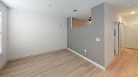 This contemporary single-floor unit is designed with a clean, modern aesthetic and consistent light wood-style flooring that flows seamlessly throughout the space. The kitchen is a standout feature, equipped with a full suite of stainless steel appliances including an electric range, built-in microwave, dishwasher, and refrigerator. Rich, wood-toned cabinetry with sleek bar-pull hardware extends from floor to ceiling, offering generous storage, while light quartz-style countertops provide ample workspace. A stainless steel sink sits beneath a window fitted with blinds, allowing natural light to filter in. The open living area presents a versatile layout with light-toned walls accented by a distinctive gray feature wall, providing an architectural focal point. A pass-through opening connects the living space to adjacent areas, enhancing the open feel. Ceiling fans and recessed lighting add to the functional design. The bathroom is thoughtfully appointed with a full soaking tub and shower combo, a wall-mounted towel bar, and a streamlined vanity featuring the same dark wood cabinetry as the kitchen, topped with a white countertop and undermount sink. A large mirror completes the bathroom's clean, cohesive look. Overall, the unit offers a modern, well-coordinated interior with consistent finishes throughout.