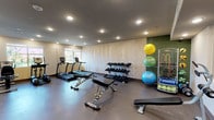 This fitness center offers a comprehensive workout environment designed to support a wide range of exercise routines. The space is finished with a neutral color palette, accented by a textured herringbone-patterned feature wall and a deep green accent wall, lending the room a modern and motivating atmosphere. Recessed lighting throughout the ceiling provides bright, even illumination across the entire floor area.

The cardio section includes treadmills positioned near a window, allowing natural light to enhance the workout experience. A multi-tier dumbbell rack stocked with a full range of free weights serves as a central feature, complemented by adjustable workout benches suitable for strength training exercises. A dedicated stretching and accessories area houses an assortment of stability balls in multiple sizes, along with resistance bands and additional training tools organized on a rack.

The flooring is a durable dark rubberized material, ideal for high-traffic fitness use and providing cushioning and safety during workouts. The layout is thoughtfully arranged to maximize open floor space, allowing for functional movement and flexibility training. Overall, this fitness center is a well-organized, fully appointed facility that caters to both cardio and strength training needs.