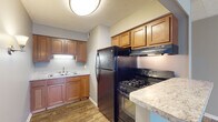 Located at 5028 Dierker Rd in Columbus, Ohio, this single-floor residence offers a practical and well-organized layout throughout. The kitchen is equipped with warm-toned wood cabinetry offering ample storage, light stone-style countertops, a double-basin sink, and black appliances including a gas range, overhead exhaust hood, and a full-size refrigerator. Wood-look flooring runs throughout the kitchen, adding a cohesive and durable finish. A hallway connects the main areas of the home, leading from the kitchen and living spaces toward the bedroom and bathroom. The living area is generously sized, featuring the same wood-look flooring, neutral gray walls, textured ceilings, and a window that allows natural light to fill the room. The bedroom is carpeted for comfort, with neutral taupe walls and a window that provides additional natural light and outdoor views. The bathroom is clean and straightforward, featuring wood-look tile flooring, a tub-shower combination with white tile surround, and a toilet, all set against gray walls. The unit's consistent neutral color palette and wood-look flooring throughout the main areas create a unified, move-in-ready aesthetic.