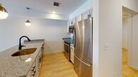 This well-appointed single-floor space offers a clean, modern aesthetic defined by neutral white walls, light wood-look flooring in the main living areas, and an abundance of natural light. The kitchen is thoughtfully designed with white shaker-style cabinetry, granite countertops, and a full suite of stainless steel appliances including a refrigerator, electric range, and over-the-range microwave — all framed by a practical galley-style layout that maximizes efficiency. Adjacent to the kitchen, the main living area features warm wood flooring and a sliding glass door with vertical blinds that opens to a private balcony, offering views of the surrounding area and filling the space with natural light. The bedrooms are finished with plush carpeting and include windows with blinds, providing a quiet and comfortable retreat. An additional flexible room features light wood flooring and a clean, versatile layout suitable for a variety of uses. The bathroom is finished with large-format tile flooring, a white vanity with granite countertop, a framed mirror, and crisp white fixtures. Throughout the space, flush-mount ceiling light fixtures provide consistent, even illumination, contributing to the overall bright and airy ambiance.