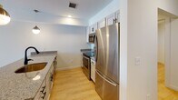 This well-appointed single-floor space offers a clean, modern aesthetic defined by neutral white walls, light wood-look flooring in the main living areas, and an abundance of natural light. The kitchen is thoughtfully designed with white shaker-style cabinetry, granite countertops, and a full suite of stainless steel appliances including a refrigerator, electric range, and over-the-range microwave — all framed by a practical galley-style layout that maximizes efficiency. Adjacent to the kitchen, the main living area features warm wood flooring and a sliding glass door with vertical blinds that opens to a private balcony, offering views of the surrounding area and filling the space with natural light. The bedrooms are finished with plush carpeting and include windows with blinds, providing a quiet and comfortable retreat. An additional flexible room features light wood flooring and a clean, versatile layout suitable for a variety of uses. The bathroom is finished with large-format tile flooring, a white vanity with granite countertop, a framed mirror, and crisp white fixtures. Throughout the space, flush-mount ceiling light fixtures provide consistent, even illumination, contributing to the overall bright and airy ambiance.