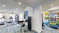 This fitness center is designed to support a comprehensive workout experience within a single-floor layout. The space features vaulted ceilings that create an open, airy atmosphere, complemented by recessed lighting and natural light streaming in through windows along the walls. The flooring features a distinctive blue and white patterned carpet that adds a modern, energetic feel to the environment.

The main workout area is equipped with a variety of strength-training machines, including cable and resistance equipment, providing users with multiple options for targeted muscle training. A dedicated stretch and accessory zone is visible to one side, stocked with stability balls in multiple sizes — yellow, green, and blue — as well as an adjustable workout bench, ideal for flexibility and supplemental training. Colorful wall art adds a vibrant touch to the otherwise clean, white-painted interior.

The layout is thoughtfully organized, with structural columns defining distinct workout zones without obstructing the overall flow of the space. The crisp white walls and bright lighting ensure excellent visibility throughout, making the fitness center feel both functional and welcoming. This space is well-suited for a range of fitness routines, from strength training to stretching and core work.