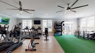 This fitness center is a comprehensively outfitted single-floor facility designed to support a wide range of workout routines. The space is finished with light wood-style flooring that transitions into green artificial turf in the dedicated training and strength zones, creating a clear visual distinction between functional areas.

The cardio section is lined with multiple treadmills, upright and recumbent stationary bikes, elliptical trainers, and a stair climber — all from True Fitness. Multiple wall-mounted flat-screen televisions are positioned throughout, and ceiling fans ensure comfortable air circulation during workouts.

The strength and training area features a multi-functional cable machine, a power rack, a freestanding heavy bag labeled "Sparring Partner," and a well-stocked dumbbell rack with a full range of weights. Plyo boxes and weight plates round out the free-weight section. A designated "Training Zone" rack offers resistance bands, battle ropes, and suspension trainers for functional fitness.

Large white-trimmed windows line multiple walls, flooding the space with natural light and offering outdoor views of the surrounding community. Full-length mirrors expand the sense of space and support form-checking during lifts. French doors provide access between zones and lead to an exterior area. Recessed lighting and crown molding add a polished, refined finish to the overall environment.