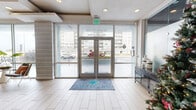 This Cortland leasing center spans a generously sized single floor, thoughtfully designed to balance professional functionality with a welcoming, contemporary aesthetic. The entry foyer greets visitors with floor-to-ceiling glass walls and double glass doors that flood the space with natural light and provide clear sightlines to the surrounding exterior. Light wood-look tile flooring runs throughout, lending warmth and visual continuity across all areas.

The main workspace features an open-plan layout with a teal accent wall, floating shelves displaying curated décor, and a central service counter with bar-height seating. Recessed lighting and exposed ductwork on a dark ceiling add an industrial-modern contrast to the otherwise bright interior. A glass-enclosed private office or conference room provides a sleek, semi-transparent meeting space visible from the main floor.

A dedicated fitness area is outfitted with rows of treadmills and strength-training equipment, dark carpeted flooring, exposed ceiling ductwork, and wall-mounted televisions — designed for practical, high-use functionality. Throughout the space, curated artwork reinforces a coastal-inspired identity, contributing to a polished, cohesive atmosphere that is both professional and approachable.