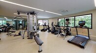 Located at 1443 Beacon St in Brookline, MA, this single-floor fitness center offers a thoughtfully designed space for comprehensive exercise and relaxation. The main workout area is generously sized, featuring durable speckled flooring and linear recessed lighting that creates a bright, energizing atmosphere. Large windows line multiple walls, flooding the space with natural light and providing views of the surrounding greenery and outdoor areas, including a glimpse of an adjacent outdoor recreation space.

The fitness floor is well-appointed with a wide range of equipment, including treadmills, ellipticals, stationary bikes, and a multi-station cable machine, along with free weights, adjustable benches, and medicine balls, accommodating both cardio and strength training needs. A wall-mounted flat-screen television and analog clock add practical touches to the workout environment.

Adjacent to the main gym floor is a welcoming lounge and refreshment area, distinguished by warm hardwood flooring, skylight panels, and a decorative geometric-tiled accent wall with subtle backlighting. This space includes a kitchenette with wood cabinetry, a round communal table with seating, and a built-in water fountain station, making it an ideal spot to rehydrate and unwind after a workout.