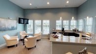 This single-floor community space is thoughtfully designed to offer a warm, inviting atmosphere across several interconnected areas. The entrance greets visitors with soaring ceilings, a striking chandelier, and a grand dark-wood front door with glass panels that flood the foyer with natural light. Neutral tones, plush carpeting, and carefully curated furnishings establish an upscale yet approachable ambiance throughout.

The main lounge area features a dramatic stone fireplace with a rustic wood mantle that reaches toward the high ceilings, serving as a focal point of the space. French doors flank the fireplace, providing direct access to an outdoor area and allowing natural light to pour in. Comfortable seating arrangements, including sofas and wingback chairs in soft, neutral tones accented with aqua and gold pillows, encourage relaxed conversation.

A separate lounge area painted in a soothing slate blue houses additional seating with cream accent chairs, a wall-mounted television, and a row of large windows offering views of an outdoor space. Adjacent to this area is a well-appointed kitchenette featuring granite countertops, pendant lighting, dark cabinetry, and a coffee station stocked for guests. The cohesive design language — black-trimmed doors, soft carpeting, and layered lighting — unifies the entire space into a polished, functional gathering environment.