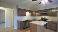 This single-story space centers around a thoughtfully designed kitchen that serves as its standout feature. The kitchen is equipped with a full suite of stainless steel appliances, including a dishwasher, electric range, and built-in microwave, providing a modern and practical cooking environment. Rich, dark wood cabinetry lines the walls in an L-shaped configuration, offering generous storage throughout. The cabinetry is complemented by light-toned countertops that create a pleasing contrast and provide ample prep space. A central island extends the counter space further, making it ideal for meal preparation or casual dining. The kitchen also features an undermount sink positioned beneath upper cabinets, with additional task lighting installed beneath them. The flooring throughout is warm-toned hardwood, adding to the welcoming atmosphere of the space. A ceiling fan with an integrated light fixture is installed overhead, contributing to both air circulation and ambient lighting. The textured ceiling adds a subtle architectural detail. An open doorway visible to the side suggests a well-connected, flowing floor plan throughout the remainder of the space.
