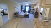This single-story residence features an open-concept layout that connects the living area to what appears to be a kitchen or dining space. The living room is generously sized, finished with neutral wall-to-wall carpeting that creates a warm, cohesive feel throughout. Crown molding lines the ceiling, adding a refined architectural detail to the space. A ceiling fan with a light fixture is centrally positioned, providing both air circulation and ambient lighting.

The living area is anchored by a large sectional sofa and paired with an upholstered ottoman, offering ample seating. A dark accent rug defines the seating zone, while an armchair adds additional comfort nearby. Decorative table lamps and wall art contribute to the layered, welcoming ambiance. A window dressed with dark curtains allows natural light to filter into the room.

Adjacent to the living area, the transition space features a warm terracotta-toned accent wall and a dark wood console, complemented by granite or stone countertop surfaces visible in the foreground, suggesting a well-appointed kitchen nearby. The overall atmosphere is comfortable, stylish, and functional.