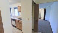 This single-floor residential space offers a practical and efficiently arranged layout designed for comfortable everyday living. The kitchen is equipped with warm-toned wooden cabinetry, a double-basin sink, and a built-in dishwasher, all complemented by light countertops and tile flooring. A window above the sink allows natural light to brighten the workspace, creating an inviting atmosphere. The kitchen flows seamlessly into the adjacent living areas through an open passageway, making the space feel connected and easy to navigate. Dark wood trim accents the doorways and baseboards throughout, providing a classic contrast against the neutral white walls. The adjoining rooms feature wall-to-wall carpeting in a neutral tone, contributing to a cozy and comfortable feel underfoot. A hallway with a closet offers convenient storage, while additional rooms visible in the background suggest a well-rounded floor plan with dedicated spaces for rest and relaxation. The overall design is clean, understated, and functional, making it well-suited for those seeking a straightforward and low-maintenance living environment on a single level.