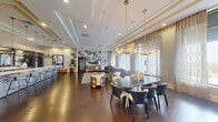 This refined amenity lounge offers a generously proportioned, single-level layout designed for both social gatherings and relaxed retreat. Rich dark hardwood flooring runs throughout, anchoring an interior defined by elevated finishes and thoughtful design details. The main gathering area features coffered and tray ceilings accented with gold trim, complemented by striking contemporary chandeliers with brass and frosted globe fixtures that cast a warm, inviting glow. A bold black-and-white abstract wall mural serves as a dramatic focal point in the lounge zone, where plush seating and a media console create a comfortable entertainment area. Floor-to-ceiling windows dressed in sheer linen curtains flood the space with natural light and frame panoramic outdoor views of the surrounding cityscape. A sophisticated dining area accommodates a large oval table with designer chairs, positioned beneath an elegant multi-globe pendant light. A marble-topped bar counter with woven rattan barstools lines one wall, offering a stylish spot for casual seating. A quiet nook tucked near the windows features two velvet armchairs and an ottoman, ideal for private conversation. The entry is marked by a statement Sputnik chandelier, setting an upscale tone from the moment one arrives. Throughout, the interplay of neutral tones, luxe textures, and curated artwork creates a cohesive and polished ambiance.