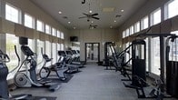 This fitness center is designed to support a comprehensive workout experience within a single-level layout. The space features soaring ceilings lined with clerestory windows along both sides, flooding the room with natural light while maintaining privacy. Two ceiling fans provide consistent air circulation throughout the facility.

The cardio zone is prominently positioned along one side and includes multiple treadmills, elliptical machines, and a stationary bike, all arranged in an orderly row. A wall-mounted television is positioned for easy viewing during cardio sessions. On the opposite side, a robust strength training area features a multi-station cable machine, a dedicated rowing machine, and additional resistance equipment, accommodating a variety of muscle-targeting exercises.

The flooring consists of durable, light-toned interlocking tiles suitable for high-traffic fitness use, with rubber mats placed beneath equipment for stability and protection. Recessed lighting supplements the natural light, ensuring the space remains well-lit at all hours. Glass-paneled double doors at the far end provide access to an adjacent area, enhancing the open, airy ambiance of the overall facility.