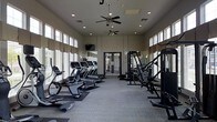 This fitness center is designed to support a comprehensive workout experience within a single-level layout. The space features soaring ceilings lined with clerestory windows along both sides, flooding the room with natural light while maintaining privacy. Two ceiling fans provide consistent air circulation throughout the facility.

The cardio zone is prominently positioned along one side and includes multiple treadmills, elliptical machines, and a stationary bike, all arranged in an orderly row. A wall-mounted television is positioned for easy viewing during cardio sessions. On the opposite side, a robust strength training area features a multi-station cable machine, a dedicated rowing machine, and additional resistance equipment, accommodating a variety of muscle-targeting exercises.

The flooring consists of durable, light-toned interlocking tiles suitable for high-traffic fitness use, with rubber mats placed beneath equipment for stability and protection. Recessed lighting supplements the natural light, ensuring the space remains well-lit at all hours. Glass-paneled double doors at the far end provide access to an adjacent area, enhancing the open, airy ambiance of the overall facility.