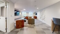 Located at 1110 Lawson Avenue in Kelowna, BC, this well-appointed amenity space offers a thoughtfully designed single-floor layout comprising a spacious lounge area, a private office or meeting room, and an additional multipurpose zone. The open living area serves as the social hub of the space, featuring plush modular seating in warm terracotta and neutral tones arranged around a central area rug. A wall-mounted flat-screen TV flanked by stylish sconce lighting anchors one wall, while a ping-pong table adds a recreational element. The room is brightened by recessed pot lighting and natural light from street-facing windows. Decorative wall art, including an oversized metallic tic-tac-toe installation and abstract sculptural pieces, lend a playful yet polished character to the space. The flooring throughout features a geometric patterned tile that adds visual interest and cohesion. The private meeting room is intimate and functional, equipped with a round marble-topped table, ergonomic seating, and a wall-mounted display screen — ideal for small group discussions or focused work sessions. A workstation area with bar-height seating provides additional productivity space. Together, these areas create a versatile, contemporary environment well-suited for both collaboration and relaxation.