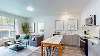 This well-designed single-floor residence offers a bright, contemporary layout that balances functionality with modern style. The open-concept living and dining area serves as the heart of the home, featuring light wood-look flooring, a comfortable sectional sofa, and a central bar-height table with stools that doubles as a casual dining space. A compact wet bar with wood-toned cabinetry and decorative wall art adds a stylish touch to this shared space. The living room is anchored by large windows that invite generous natural light and provide outdoor views, complemented by a wall-mounted TV, a sleek media console, and a ductless mini-split HVAC unit for efficient climate control. The kitchen is well-appointed with stainless steel appliances including a refrigerator, range, and microwave, alongside crisp white upper cabinets, wood-toned lower cabinets, and white quartz countertops. Two bedrooms offer restful retreats, each filled with natural light from large windows with blinds. One bedroom includes a spacious double closet. Both bathrooms feature wood-toned vanities with white countertops and hard-surface flooring. A dedicated laundry area with a stacked washer and dryer adds everyday convenience. Light wood-look flooring flows consistently throughout the entire space, reinforcing its clean, cohesive aesthetic.