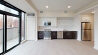 This single-floor space showcases a clean, modern design with an open-concept layout and light hardwood-style flooring throughout the main living areas. The kitchen is thoughtfully appointed with two-toned cabinetry — dark espresso lower cabinets paired with crisp white upper cabinets — complemented by white quartz countertops and a classic subway tile backsplash. Stainless steel appliances, including a gas range, microwave, dishwasher, and refrigerator, are integrated seamlessly. Open floating shelves add both function and visual interest to the kitchen wall.

The spacious living area is bathed in natural light through large floor-to-ceiling windows and sliding glass doors that open directly onto a private balcony, offering urban views. A ceiling fan enhances comfort in this generously proportioned space. Sliding barn-style closet doors lead to well-organized storage areas with built-in shelving and hanging rods.

The bathroom maintains the unit's modern aesthetic with dark large-format floor tiles, a wall-mounted floating vanity with a white surface and rectangular undermount sink, a frameless glass walk-in shower with tile surround, and recessed lighting. The overall ambiance is bright, minimal, and contemporary throughout.