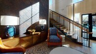 Located at 275 Huger St in Charleston, South Carolina, this thoughtfully designed single-floor amenity space blends contemporary aesthetics with warm, curated details to create a welcoming communal environment. The grand lobby-style lounge immediately impresses with soaring double-height ceilings, a sculptural staircase with dark metal railings and wood treads, and a dramatic textured feature wall finished in rich, geometric metallic panels. Layered lighting, bold artwork, and a mix of leather and fabric seating lend an inviting, upscale character. A generously proportioned living lounge area offers oversized modular seating, a large-screen television, and abstract wall art, bathed in natural light from large windows with views of the surrounding greenery. A striking floor-to-ceiling wood-and-teal shelving unit anchors an adjacent seating nook, adding a library-like warmth to the space. The kitchen area is finished with sleek grey cabinetry accented by gold hardware, marble-look countertops, a bold blue subway tile backsplash, and built-in stainless steel appliances. The dining area features a solid wood communal table, mid-century style wooden chairs, and eye-catching geometric-patterned flooring in blue and grey tones, beneath statement brass pendant lights. Glass doors open to an outdoor area, filling the space with abundant natural light.