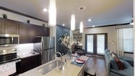 This thoughtfully designed single-floor residence showcases a modern open-concept layout that seamlessly connects the kitchen, dining, and living areas. The kitchen is a standout feature, centered around a spacious island with light stone countertops, an undermount sink, and a sleek faucet — ideal for both meal preparation and casual dining. Dark espresso cabinetry provides a bold contrast against the lighter countertops, while stainless steel appliances, including a full-size refrigerator, add a contemporary touch. Three elegant cylindrical pendant lights hang above the island, creating a warm and inviting focal point. Recessed lighting throughout ensures the space remains bright and well-illuminated. A striking teal accent wall adds a pop of color and visual depth, complementing the rich hardwood flooring that runs throughout. A built-in shelving unit offers functional display space, while a dedicated workspace nook with a desk and chair provides practicality. Natural light filters in through a glass-paneled door fitted with blinds, connecting the interior to the outside. The overall ambiance is modern, warm, and functional — a well-appointed space designed for comfortable everyday living.