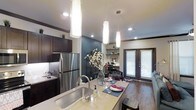This thoughtfully designed single-floor residence showcases a modern open-concept layout that seamlessly connects the kitchen, dining, and living areas. The kitchen is a standout feature, centered around a spacious island with light stone countertops, an undermount sink, and a sleek faucet — ideal for both meal preparation and casual dining. Dark espresso cabinetry provides a bold contrast against the lighter countertops, while stainless steel appliances, including a full-size refrigerator, add a contemporary touch. Three elegant cylindrical pendant lights hang above the island, creating a warm and inviting focal point. Recessed lighting throughout ensures the space remains bright and well-illuminated. A striking teal accent wall adds a pop of color and visual depth, complementing the rich hardwood flooring that runs throughout. A built-in shelving unit offers functional display space, while a dedicated workspace nook with a desk and chair provides practicality. Natural light filters in through a glass-paneled door fitted with blinds, connecting the interior to the outside. The overall ambiance is modern, warm, and functional — a well-appointed space designed for comfortable everyday living.
