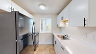 This single-floor unit accommodates three bedrooms, one bathroom, a kitchen, and a living room within a efficiently organized footprint. Throughout the unit, a consistent neutral color palette of white and beige tones creates a clean, cohesive aesthetic, complemented by textured ceilings and plush carpeting in the living and sleeping areas.

The living room is a bright, open space anchored by a sliding glass door that opens to a balcony, offering outdoor views and natural light. The galley-style kitchen features white shaker cabinets with matte black hardware, white countertops, a stainless steel sink, and black appliances including a refrigerator and range. A window above the counter provides additional natural light. The flooring here is durable tile in a warm stone tone.

The primary bedroom is a well-proportioned room with a window overlooking the exterior. The two additional bedrooms are carpeted and offer flexible use. One bedroom includes a sliding-door closet with built-in hanging rods. The bathroom features a full tub-shower combination, a white vanity with cabinetry, a toilet, and tile flooring matching the kitchen.