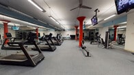 Located at 1349 N North Park Ave in Chicago's Lincoln Park neighborhood, this fitness center occupies a single-floor space designed to accommodate a comprehensive range of workout activities. The open floor plan provides ample room for members to move freely between equipment zones without feeling crowded.

The space is defined visually by its bold red structural columns, which extend from floor to ceiling and serve as striking architectural focal points throughout the facility. The flooring consists of durable gray rubberized material suited for heavy equipment use. Linear fluorescent lighting runs across the ceiling, providing consistent, even illumination throughout the space. Ceiling fans are mounted overhead to aid in air circulation.

Large mirrors line multiple walls, creating a sense of expanded depth and allowing users to monitor their form during exercise. The cardio section is well-stocked with Matrix treadmills, ellipticals, stationary bikes, and rowing machines arranged in organized rows. The strength training area features cable machines, weight benches, a dumbbell rack, and plate-loaded equipment. Multiple flat-screen televisions are mounted from the ceiling throughout the space, offering entertainment during workouts. A separate windowed room visible from the main floor appears to provide additional training space.