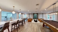 Located on Hickory Club Drive in Nashville, TN, this single-floor community amenity space is thoughtfully designed for socializing and recreation. The open-concept layout seamlessly connects a well-appointed kitchen, a dining bar area, and an expansive lounge and game room.

The kitchen features white shaker cabinetry with black hardware, white quartz countertops, stainless steel appliances, and ample natural light from a large window. It connects directly to a central island bar fitted with a sink, gold-toned faucet, and barstool seating beneath a row of modern globe pendant lights with brass accents — an inviting hub for casual gatherings.

The living area is anchored by a curated seating arrangement of cognac leather armchairs around a round black coffee table, set against an accent wall in charcoal gray adorned with a bold teal and green abstract artwork. Sheer floor-length curtains line the perimeter walls, softening the abundant natural light that pours in through multiple windows and glass French doors leading to an outdoor patio.

A dedicated game zone includes a full-size billiards table and a shuffleboard table, while a striking slatted dark-wood feature wall hosts dual flat-screen televisions, creating a stylish entertainment focal point. Light wood-look flooring flows consistently throughout, tying the entire space together with a warm, contemporary aesthetic.
