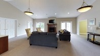 This single-floor clubhouse is thoughtfully designed to offer a variety of social and functional spaces for its members. The main lounge serves as the heart of the facility, boasting soaring vaulted ceilings, plush carpeting, and an abundance of natural light streaming through multiple windows and French doors. A traditional wood-mantled fireplace anchors the room, accompanied by a wall-mounted television, comfortable dark upholstered sofas, and accent chairs in warm patterned fabrics. Adjacent to the lounge is a combined billiards and library room, distinguished by rich dark wood crown molding, built-in bookshelves filled with reading material, and a full-size pool table beneath a pendant light. Accent seating with chevron-patterned upholstery adds a stylish touch. A separate dining area features a round table with dark chairs and connects seamlessly to a wet bar with bar-height seating, making it well-suited for social gatherings. A dedicated computer room provides a quiet workspace with a desk, computer setup, and natural light from a window. Throughout the clubhouse, neutral warm-toned walls and carpeted floors create a cohesive and inviting atmosphere ideal for both relaxation and recreation.