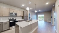 This single-story residence is thoughtfully designed with a contemporary aesthetic, combining modern finishes with functional living spaces throughout. The open-concept kitchen serves as the heart of the home, anchored by a generous center island topped with striking white quartz countertops and equipped with an undermount sink and matte black faucet. Two globe pendant lights hang above the island, complementing the recessed lighting overhead. Two-toned cabinetry pairs lighter upper cabinets with darker lower units, while stainless steel appliances, including a refrigerator, electric range, and microwave, complete the well-appointed cooking space. Light wood-look flooring extends seamlessly from the kitchen into the living area, which features a sliding glass door opening to a private balcony overlooking a natural outdoor setting.

The space includes two bedrooms, each carpeted and filled with natural light from large windows. The primary bedroom connects to an en suite bathroom featuring a sliding barn door entry, wood-toned vanity with white countertop, matte black fixtures, and light vinyl flooring. A second full bathroom offers a tub-shower combination with matching finishes. A dedicated walk-in closet with wire shelving provides ample storage, and an in-unit laundry room houses a full-sized washer and dryer.