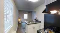 This single-floor space offers a functional and modern layout centered around an open-concept design that seamlessly connects the kitchen and living area. The kitchen is well-appointed with white shaker-style cabinetry, dark countertops, a stainless steel double-basin sink, and black appliances including a range and refrigerator. An overhead range hood provides practical ventilation. The peninsula-style counter serves as a natural divider between the kitchen and the adjoining living area, enhancing the open feel of the space. The living area features wood-style plank flooring that runs throughout, adding warmth and visual continuity. A ceiling fan with a built-in light fixture is centrally installed, promoting air circulation and ambient lighting. Natural light filters in through multiple windows fitted with blinds, creating a bright and welcoming atmosphere. The neutral gray wall tones complement the flooring and cabinetry, lending the space a cohesive and contemporary aesthetic. The textured ceiling adds a subtle design element overhead. Overall, the layout is efficient and well-designed, making thoughtful use of the available space while maintaining a clean, modern look throughout.