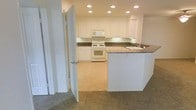 This single-story space is designed with a clean, functional layout that centers around a well-appointed kitchen. The kitchen is outfitted with crisp white cabinetry that extends to the upper walls, providing generous storage space. Dark granite-style countertops offer a striking contrast against the white surfaces, while a built-in peninsula with a sink serves as a natural dividing point between the kitchen and the adjoining carpeted living area. Recessed lighting is installed throughout the ceiling, ensuring the space is evenly and thoroughly illuminated. White appliances, including a range and over-the-range microwave, are seamlessly integrated into the cabinetry. The kitchen floor features smooth, light-toned vinyl or tile, transitioning to carpet in the adjacent living space, creating a clear and practical delineation between functional and relaxation zones. A doorway visible from the kitchen provides access to additional areas of the home. The overall aesthetic is neutral and bright, with a cohesive color palette of whites and warm tones that lends the space a tidy, welcoming ambiance suited for everyday living.