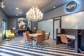 Located at 1205 Maple St in Carrollton, Georgia, The Jim is a single-floor commercial space that blends bold design with functional versatility. The space is immediately defined by its striking black-and-white diagonal striped tile flooring, which flows throughout the entire interior and establishes a dramatic visual rhythm underfoot.

Teal trim and door frames frame each zone, creating strong color contrast against the cool gray walls and ceilings. Crystal chandeliers suspended throughout the space add a glamorous, upscale touch that balances the space's graphic, modern aesthetic.

A dedicated workstation area features sleek dark cabinetry and ergonomic rolling chairs arranged around individual desks, while a glowing circular neon sign reading "The Jim" serves as a bold focal point. Adjacent to this area is a compact kitchenette outfitted with a coffee station, sink, and under-counter cabinetry — ideal for serving clients.

A business center section offers a bar-height counter with red upholstered stools and multiple computer monitors, bathed in natural light from large glass doors that open to an outdoor area. A lounge zone invites relaxation with a plush navy velvet sofa, vibrant yellow accent chairs, gold decorative lamps, and a large mirrored wall panel that amplifies the sense of depth and light throughout the space.
