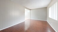 Located at 12603 Eucalyptus Ave in Hawthorne, California, this single-story, two-bedroom unit offers a practical and well-organized layout. Rich, warm-toned hardwood-style flooring flows throughout the living, dining, and bedroom areas, creating a cohesive and inviting atmosphere. The living room features a textured ceiling, vertical blind-covered windows that allow natural light to filter in, and distinctive vertical wood panel wall accents. It opens into a combined dining area, which transitions into the kitchen. The kitchen is equipped with white cabinetry, white countertops, a gas range, a stainless steel refrigerator, and a range hood, all set atop light-toned tile flooring. A large window in the dining area further brightens the space. Both bedrooms continue the warm hardwood-style flooring and are fitted with ceiling fans and windows with vertical blinds. The primary bedroom includes a walk-in closet, while the second bedroom offers a sliding-door closet. A hallway connects the bedrooms to the main living areas, providing a clear and efficient flow throughout the unit. Neutral gray walls throughout lend a clean, move-in-ready aesthetic to this Hawthorne residence.