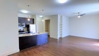 This single-floor space showcases an open-concept layout that seamlessly connects the kitchen and living areas, creating a cohesive and functional environment. The kitchen is equipped with stainless steel appliances, including a refrigerator, stove, and microwave, complemented by dark cabinetry and a granite-topped island that doubles as a breakfast bar. Pendant lighting hangs above the kitchen area, adding focused illumination and a contemporary design touch. The backsplash adds subtle texture to the cooking space.

Adjacent to the kitchen, a walk-in storage or pantry area provides generous organizational space with multiple built-in shelves. A built-in shelving unit along one wall of the living area offers additional storage and display opportunities. The open living space is generously proportioned, allowing for flexible furniture arrangements. Warm hardwood flooring runs throughout the entire unit, contributing to a cohesive and inviting atmosphere. Flush ceiling lights ensure consistent, even illumination across the space. The neutral wall tones and clean architectural lines give the interior a modern, understated aesthetic.