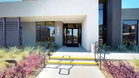 This striking single-story commercial space makes an immediate impression with its modern, desert-contemporary architecture. The exterior entrance is clean and minimalist, framed by desert landscaping, large dark-framed glass doors, and a covered entryway that sets the tone for the refined interior beyond.

Inside, the open lobby and lounge area commands attention with soaring double-height ceilings and expansive floor-to-ceiling glass curtain walls that flood the space with abundant natural light and offer views of the surrounding outdoor landscape, including mature palm trees. A towering double-sided mosaic stone fireplace serves as the dramatic centerpiece of the room, rising from floor to ceiling and anchoring the seating area with bold architectural presence.

The lounge is thoughtfully furnished with low-profile sectional sofas in charcoal and accents of deep plum, complemented by organic wood stump side tables and a textured area rug that grounds the seating arrangement. Sleek cylindrical pendant lighting descends from the ceiling, adding a contemporary touch. To one side, a glass-walled office or private meeting room with frosted privacy panels provides a functional counterpoint to the open common area. Light-toned large-format tile flooring runs throughout, enhancing the sense of spaciousness and continuity across the entire floor.