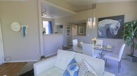 This single-floor residence showcases an open-concept design that connects the living, dining, and kitchen areas into one cohesive space. The living area is anchored by a white tufted sofa accented with blue and grey decorative pillows, complemented by small potted plants that add a touch of greenery. A large abstract artwork adorns the wall, adding a bold visual focal point to the room.

The dining area features a glass-top table surrounded by white chairs, set beneath a striking cascading crystal and chrome chandelier that serves as a standout design element. A fresh bouquet of yellow tulips on the table adds a warm, inviting touch.

A half-wall pass-through connects the kitchen to the main living space, offering both functional visibility and an open feel. The kitchen is partially visible beyond, where a window dressed with blue curtains allows natural light to filter through.

Neutral warm-beige tones coat the walls throughout, contributing to a calm and cohesive aesthetic. The space benefits from recessed and track lighting, ensuring consistent illumination. Hallways leading to additional rooms are visible, hinting at a thoughtfully laid-out floor plan throughout the home.