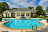 Located in Sandy Springs, Georgia, this single-level outdoor amenity space at 100 Highland Park Trail offers a well-appointed retreat designed for relaxation and recreation. At its centerpiece is a generously sized, freeform swimming pool with crystal-clear blue water, smooth concrete coping, and entry handrails. The pool is complemented by a classic clubhouse building featuring a covered porch with white columns, stone accents, a pitched roof with a chimney, and large windows that overlook the water.

The expansive pool deck is lined with a row of sling-style chaise lounges, providing ample space for sunbathing, while shaded umbrella seating areas offer a cooler alternative for lounging. Large decorative planters with colorful flowers add vibrant touches throughout the pool surround.

Beyond the pool area, a secondary outdoor patio offers intimate seating with wrought-iron tables and chairs beneath blue market umbrellas, ideal for casual gatherings. The space is enclosed by decorative black iron fencing and a keyed entry gate, ensuring privacy and security. A pergola structure adds architectural interest near the entrance. Mature trees and meticulously maintained hedgerows frame the entire amenity area, creating a lush, park-like atmosphere throughout.