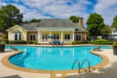 Located in Sandy Springs, Georgia, this single-level outdoor amenity space at 100 Highland Park Trail offers a well-appointed retreat designed for relaxation and recreation. At its centerpiece is a generously sized, freeform swimming pool with crystal-clear blue water, smooth concrete coping, and entry handrails. The pool is complemented by a classic clubhouse building featuring a covered porch with white columns, stone accents, a pitched roof with a chimney, and large windows that overlook the water.

The expansive pool deck is lined with a row of sling-style chaise lounges, providing ample space for sunbathing, while shaded umbrella seating areas offer a cooler alternative for lounging. Large decorative planters with colorful flowers add vibrant touches throughout the pool surround.

Beyond the pool area, a secondary outdoor patio offers intimate seating with wrought-iron tables and chairs beneath blue market umbrellas, ideal for casual gatherings. The space is enclosed by decorative black iron fencing and a keyed entry gate, ensuring privacy and security. A pergola structure adds architectural interest near the entrance. Mature trees and meticulously maintained hedgerows frame the entire amenity area, creating a lush, park-like atmosphere throughout.