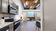 Located at 1100 N 2nd St in Leavenworth, Kansas, this one-bedroom, one-bathroom loft masterfully blends industrial character with contemporary design. The open-concept layout connects the kitchen, living, and sleeping areas, all unified by warm hardwood-style flooring and soaring ceilings adorned with original wood beams and exposed ductwork. A ceiling fan complements the dramatic overhead lighting, while the structural wood ceiling adds rich texture and warmth to the space.

The kitchen features stainless steel appliances, including a range, microwave, and refrigerator, paired with sleek gray cabinetry. A barn-style sliding door with frosted glass panels separates the living area from the bedroom, offering privacy without sacrificing the loft's open feel. The living area showcases a rustic original support column, adding an authentic industrial touch.

The generously sized bedroom is highlighted by floor-to-ceiling windows framed by exposed brick walls, flooding the space with natural light while maintaining an intimate atmosphere. A large closet provides ample storage.

The bathroom is clean and well-appointed, featuring a full tub/shower combo, a granite-topped vanity with an undermount sink, and hardwood-style flooring that ties seamlessly into the rest of the unit.