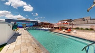 Located at 947 N Park Ave in Tucson, Arizona, this expansive rooftop amenity deck offers a thoughtfully designed outdoor retreat with a wide range of leisure and social features. The centerpiece is a large, sparkling resort-style pool with a tiled mosaic accent wall and an adjacent spa, surrounded by lounge chairs and ample deck space finished in warm-toned stone pavers. A vibrant mural adds an artistic focal point to the poolside atmosphere.

The rooftop is divided into distinct zones, each serving a different purpose. A dedicated lounge area features red-canopied pergolas and turf lawn seating, providing shaded relaxation space. String lights suspended between modern steel posts illuminate an open-air gathering and dining area, furnished with bar-height seating and outfitted with raised concrete planter beds filled with native desert grasses and flowering plants. An outdoor fireplace creates a cozy focal point for evening gatherings.

Lush landscaping throughout includes towering cacti, ornamental grasses, purple ground cover, and desert-adapted plantings that reflect the Sonoran surroundings. The rooftop offers panoramic views of the Tucson mountain ranges and the open Arizona sky, creating a dramatic backdrop for the entire space. The design seamlessly blends urban sophistication with regional desert character.