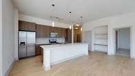 This single-floor space showcases a thoughtfully designed open-concept layout that seamlessly connects the kitchen and living areas. The kitchen is a standout feature, anchored by a generously sized central island with a crisp white countertop and an integrated sink, providing ample workspace and a natural gathering point. Full-height dark wood cabinetry lines the kitchen walls, offering abundant storage while adding warmth and contrast to the light-toned space. A classic subway tile backsplash runs beneath the upper cabinets, complementing the stainless steel appliance suite that includes a refrigerator, microwave, and range. Pendant lighting and a track light fixture hang above the kitchen, providing focused illumination throughout the area. Light wood-style flooring flows continuously across the open floor plan, enhancing the sense of spaciousness and continuity. The adjacent living area features a built-in shelving niche with a lower ledge, offering a versatile space for display or storage. White walls and clean architectural lines throughout contribute to a bright, contemporary ambiance. The open sightlines between rooms create an inviting, connected atmosphere well-suited for both everyday living and entertaining.