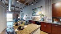This single-floor urban loft masterfully blends industrial character with contemporary design. The open-concept living and kitchen area is anchored by soaring exposed concrete ceilings with visible steel beams, ductwork, and track lighting, all complemented by raw brick accent walls that lend an authentic warehouse aesthetic. Polished concrete floors run throughout, unifying each space with a sleek, modern continuity.

The kitchen is well-appointed with rich wood cabinetry, granite countertops, a tile backsplash, and black appliances including a refrigerator and dishwasher. A large central island with seating provides additional prep space and serves as a natural gathering point, seamlessly connecting the kitchen to the living area.

The living room offers an inviting atmosphere, featuring bold large-format artwork, a comfortable seating arrangement, and dramatic floor-to-ceiling brick walls that emphasize the loft's generous vertical scale.

Two bedrooms provide comfortable retreats; the primary bedroom is highlighted by an oversized window framing expansive city and treetop views, along with an exposed brick feature wall and polished concrete floors. The secondary bedroom is a quiet, well-lit space.

Two bathrooms complete the layout — one with a walk-in tiled shower, the other with a tub-shower combination — both finished with granite vanities and warm wood cabinetry consistent with the overall design palette.