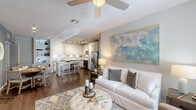 This single-floor residence is thoughtfully designed with an open-concept layout that seamlessly connects the kitchen, dining, and living areas. The kitchen is a standout feature, outfitted with white shaker cabinetry, granite countertops, a classic subway tile backsplash, and a full suite of stainless steel appliances. A granite-topped island with woven bar stools serves as both a prep space and casual dining area, while track lighting illuminates the room throughout. Adjacent to the kitchen, the dining area accommodates a round table with woven chairs and benefits from proximity to a stacked in-unit washer and dryer. The living room is bright and welcoming, featuring dark hardwood-style flooring, a ceiling fan, and a large window that allows natural light to fill the space. The bedroom is a calm retreat with neutral tones, plush carpet, natural light from a window, and ample room for full-sized furniture. The bathroom features a granite-topped vanity with grey cabinetry, light tile flooring, and a full soaking tub with tiled surround. Built-in open shelving in the living area provides additional storage and display space throughout.