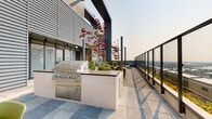 Located atop a modern building at 8460 Ash St in Vancouver, BC, this rooftop patio is a thoughtfully designed outdoor amenity space that combines functionality with striking urban scenery. The patio features a contemporary aesthetic, with a mix of large-format stone tile and warm composite wood decking that defines distinct zones across the rooftop.

At one end, a built-in outdoor kitchen island houses a stainless steel multi-burner BBQ grill, providing a dedicated space for outdoor cooking and entertaining. Adjacent to this, raised concrete planters filled with ornamental trees, grasses, and seasonal plants add a lush, garden-like atmosphere to the space.

Multiple seating and dining configurations are spread throughout the patio, including a large communal wood-topped table with green upholstered chairs set within a planted enclosure, as well as a separate dining set with a shade umbrella ideal for alfresco meals. A wood-decked lounge area with casual green furniture offers a relaxed setting for gathering.

Strung overhead with Edison-style string lighting, the patio transitions seamlessly from day to evening use. A sleek glass-and-steel perimeter railing runs along the edge of the rooftop, ensuring unobstructed panoramic views of the Vancouver skyline and surrounding cityscape. The building's corrugated metal cladding provides a modern industrial backdrop throughout the space.
