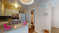 Located at 11600 Huebner Rd in San Antonio, Texas, this single-floor unit offers a practical and well-organized layout suited for comfortable everyday living. The kitchen is efficiently designed with white cabinetry, a mosaic tile backsplash, granite-look countertops, and a full suite of white appliances including a refrigerator, electric range, and dishwasher. A peninsula breakfast bar connects the kitchen to the main living area, providing additional counter space and a casual dining option. The living area is generously sized, featuring warm neutral-toned carpet, clean white baseboards, and multiple windows that allow natural light to fill the space. The open flow between the kitchen and living room creates an inviting and functional common area. A full bathroom is accessible from the main hallway and includes a shower-tub combination. A dedicated walk-in closet with hardwood-style flooring and built-in shelving offers ample storage. The unit also features central air and heating with visible vents throughout. The overall aesthetic is clean and neutral, providing a versatile backdrop adaptable to a variety of interior styles.