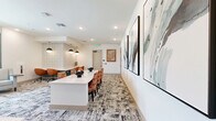 Located at 904 S Lake St in Leesburg, Florida, this thoughtfully designed single-floor workspace offers a professional and modern environment ideal for collaborative work and meetings. The space is organized into distinct functional zones, all unified by a cohesive contemporary aesthetic featuring patterned carpet, crisp white walls, and carefully curated décor.

The main open area houses two separate workstation configurations. The first is a long white quartz-topped desk set against a textured stone-accent wall, illuminated by a trio of sleek pendant lights and equipped with ergonomic burnt-orange chairs — providing an efficient, multi-user workstation. The second is a conference-style table surrounded by upholstered coral-toned chairs, with a triptych of abstract artwork adorning the white wall behind it, creating a polished meeting environment.

A relaxed lounge corner features a light gray accent chair positioned near a large window dressed with teal curtains, allowing natural light to fill the space and offering views to the outside. A built-in workstation nook with a quartz countertop, wall sconce lighting, and vibrant botanical wall art provides a more private, focused work setting.

The space also includes a bathroom and is finished with recessed ceiling lighting throughout, contributing to a bright, welcoming ambiance suited for productivity.