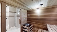 This single-level sauna facility offers a focused and functional wellness experience. The sauna room itself is lined with horizontal wood paneling across both the walls and ceiling, creating a warm, enveloping aesthetic consistent with traditional sauna design. A centrally positioned electric sauna heater with a stone top serves as the room's focal point, mounted within a protective wooden surround. Slatted wooden benches provide seating and reclining space for users during heat sessions. Overhead lighting offers a soft, ambient glow throughout the room. A glass-paneled door provides access to an adjoining changing or locker area, which is visible through the entrance and features built-in lockers along the walls and clean, light-toned flooring. The transition between the warm wood interior of the sauna and the bright, utilitarian finish of the adjacent space creates a clear functional separation. Safety and instructional notices are posted within the sauna, reflecting attention to user guidelines. Overall, the space is designed with practicality and wellness in mind, providing a clean and efficient environment suited for relaxation and heat therapy.