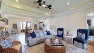 The Cottages is a thoughtfully designed single-story property that blends classic architectural details with warm, welcoming interiors. The main living area immediately draws attention with its elevated tray ceilings adorned with crisp crown molding, lending a sense of height and refinement to the space. A decorative ceiling fan anchors the room while adding both function and style. Hardwood flooring runs throughout, contributing warmth and a cohesive aesthetic across the open layout. The living area is generously proportioned, featuring a comfortable neutral sofa accented with navy blue throw pillows, a woven rattan ottoman serving as a centerpiece coffee table, and a bold navy accent chair that adds a pop of color. Recessed lighting complements the natural light that filters in through elegant French doors at the far end of the room, which open to reveal additional interior spaces beyond. Subtle architectural beams add character to the ceiling, while the soft, neutral wall palette creates a calm and airy ambiance throughout. Greenery and tasteful décor accents complete the inviting atmosphere, making The Cottages a space that feels both polished and approachable.