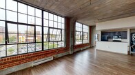 This urban loft showcases a striking industrial aesthetic defined by its raw, exposed concrete ceilings, classic red brick walls, and oversized steel-framed windows that span nearly floor to ceiling. The generous open-plan living area is flooded with natural light from multiple large window banks, framing captivating views of the surrounding cityscape and street-level activity below. Rich hardwood flooring runs throughout, providing warmth that contrasts beautifully with the rugged industrial elements overhead, including exposed ductwork and structural beams. The kitchen is equipped with dark cabinetry, black appliances including a range with microwave, and a convenient breakfast bar pass-through that connects seamlessly to the main living area. A stacked washer and dryer unit is neatly integrated into the kitchen space. The bathroom features a full tub-shower combination with chrome fixtures and accessibility grab bars. Additional rooms include private enclosed spaces accessed through white interior doors, with wall sconces providing ambient lighting. The exposed concrete ceiling and industrial piping continue throughout, maintaining a cohesive loft character across the entire single-floor layout. This space masterfully balances raw architectural character with functional modern living essentials.