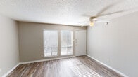 This two-floor residence offers a clean, functional layout with consistent design elements throughout. The main living area welcomes visitors through a front entry door flanked by two windows fitted with horizontal blinds, allowing natural light to cast across the light-toned wood-look flooring. A ceiling fan with an integrated light fixture is centrally positioned in this space. Adjacent rooms on this floor share the same flooring and neutral greige wall tones, with additional windows providing ample daylight. The galley-style kitchen is efficiently designed with white cabinetry lining both sides, speckled laminate countertops, a double-basin stainless steel sink, a coil-burner electric range, and a full-size refrigerator. A full bathroom features a tiled shower/tub combination and wood-look flooring. A hallway connects the rooms on the upper floor, which features darker hardwood-style flooring, continuing the warm, cohesive aesthetic. Throughout both floors, textured popcorn ceilings and white baseboards provide a consistent finish. The open corridor leads to additional rooms bathed in natural light from large windows, reinforcing a bright and airy atmosphere across the entire space.