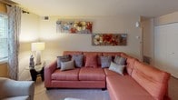 This single-floor residence is thoughtfully organized to maximize both comfort and functionality. The living room serves as the heart of the home, featuring plush carpeting and warm, neutral walls that create an inviting atmosphere. A spacious sectional sofa anchors the room, complemented by decorative wall art that adds a vibrant touch.

Adjacent to the living area, the dining space accommodates a round glass-top table with upholstered chairs, a sideboard, and a decorative mirror, with a window providing natural light. The kitchen is equipped with ample cabinetry, laminate countertops, a gas range, and tile flooring, offering a practical and efficient cooking environment.

The primary bedroom features carpeted floors, multiple windows with drape-adorned curtain rods that invite natural light, and space for a dresser and bedside furnishings. A secondary bedroom includes a dark-framed bunk bed setup with closet access, making it well-suited for shared use. The bathroom is outfitted with a vanity featuring a single-basin sink, speckled countertop, a lighted mirror, towel rack, and a shower-tub combination with a patterned curtain, completing the home's practical amenities.